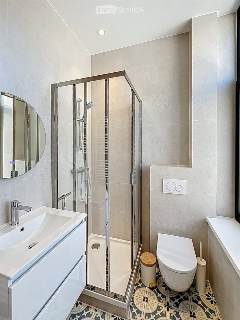 Modern bathroom with a white sink, chrome faucet, round mirror, toilet, glass-enclosed shower, and decorative tile floor.