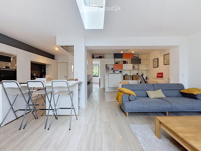 Open-plan living and dining area with a grey sofa, wooden coffee table, and four white stool chairs around a kitchen island. The kitchen features white cabinets, a microwave, and an oven. Shelves hold books and decorative items.