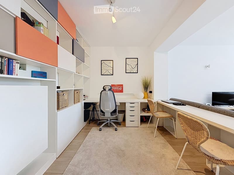 A modern, well-lit room with a desk, chair, shelves, and a small seating area with a couch and a TV.