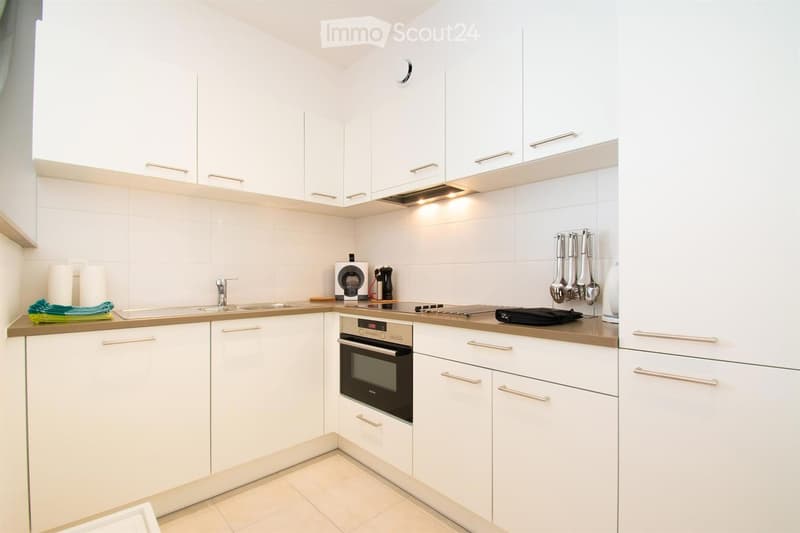 Modern kitchen with white cabinets, stainless steel appliances, and a light-colored countertop. Features include a sink, faucet, oven, coffee maker, and oven mitts.