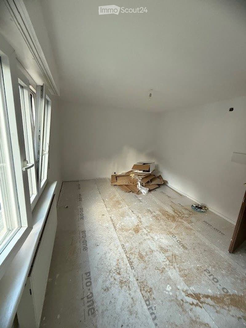 Empty room with white walls, wooden floor, and a window. Some cardboard boxes and debris are present on the floor.