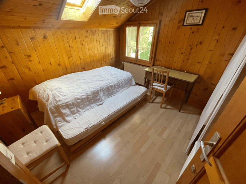 Cozy attic bedroom with wooden walls and floors, featuring a single bed, a small desk with a chair, and a cushioned stool near the bed.