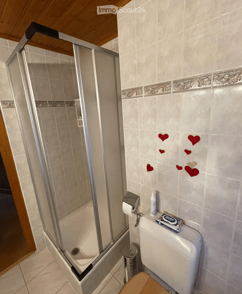 Bathroom with a glass-enclosed shower, toilet, toilet paper holder, a small bottle on the toilet tank, and a wooden stool in front of the toilet. The walls are tiled with decorative borders and small red heart stickers.