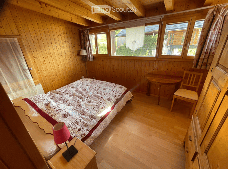 Cozy wooden bedroom with a bed, nightstand with lamp, small table and chair, and large windows with curtains.