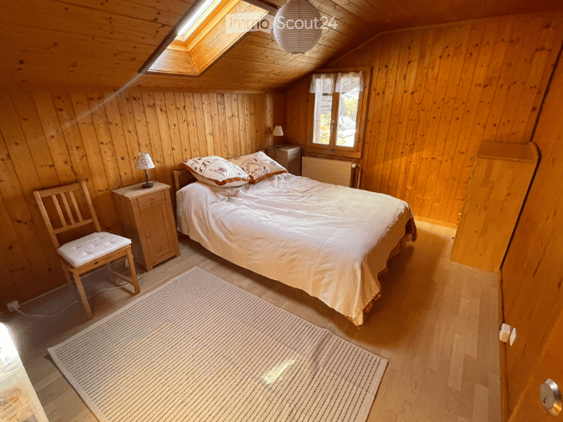 Cozy wooden bedroom with slanted ceiling, skylight, bed with pillows and blanket, nightstand with lamp, wooden chair, and radiator.