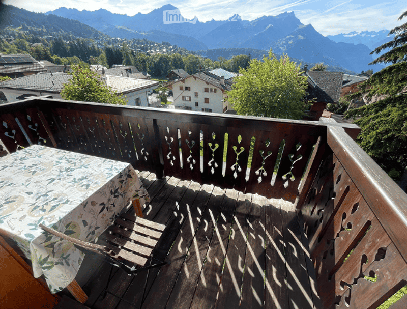 Balcony with wooden decking, decorative railing, and a table with a floral tablecloth, overlooking a scenic mountain valley.
