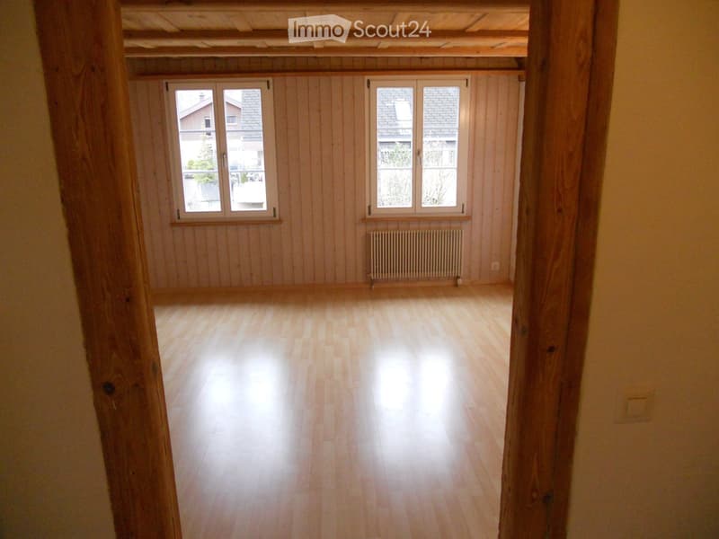 Empty room with wooden flooring, light wooden walls, and large windows with white frames. A radiator is visible under one window.
