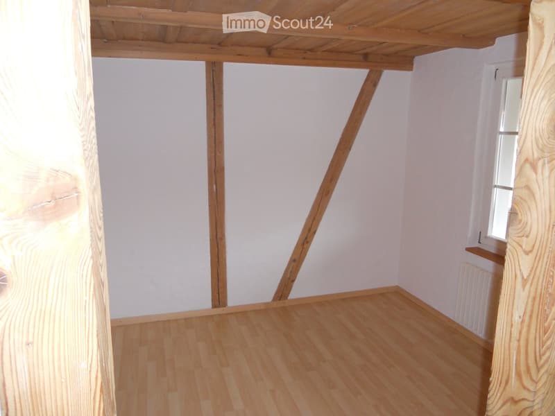 Empty room with wooden flooring, white walls, and exposed wooden beams on the ceiling. A small window is visible on one wall.