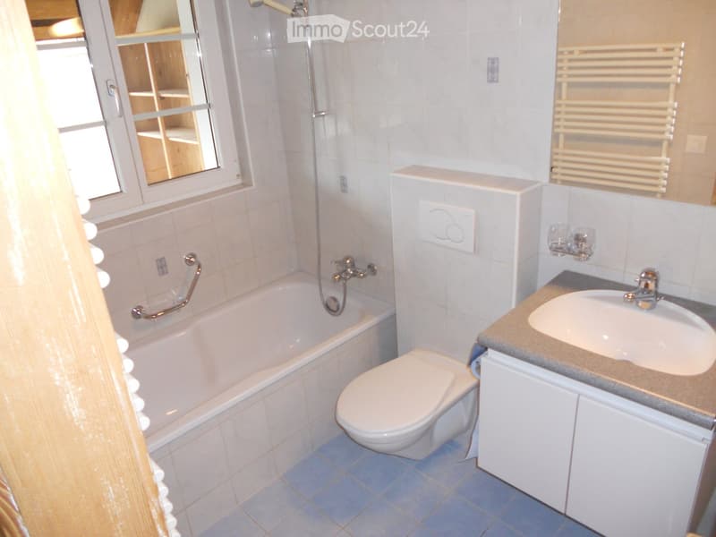 Modern bathroom with a bathtub, toilet, sink, and a mirror above the sink. The room features tiled walls and a radiator on the right side.