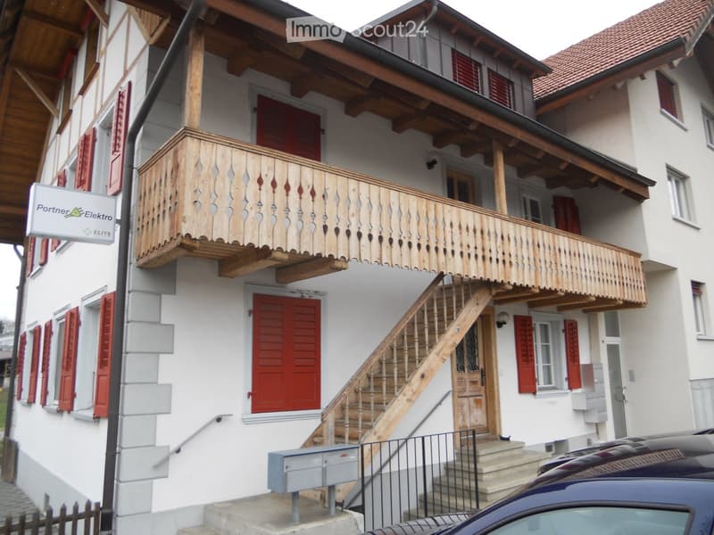 Two-story residential building with white walls, red shutters, and a wooden balcony on the upper floor. A wooden staircase leads to the entrance, and a car is parked in front.