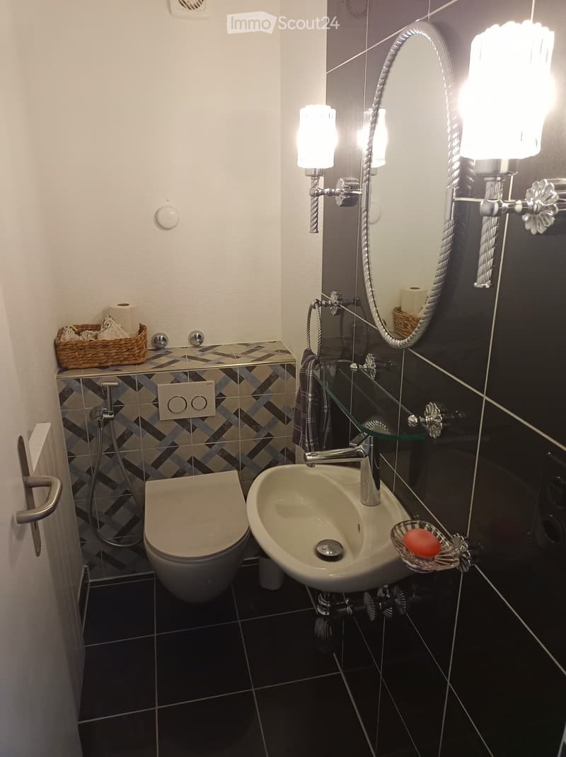 Modern bathroom with dark tiled walls, a toilet, a sink with a chrome faucet, a large mirror with built-in lighting, and a wicker basket on the counter.
