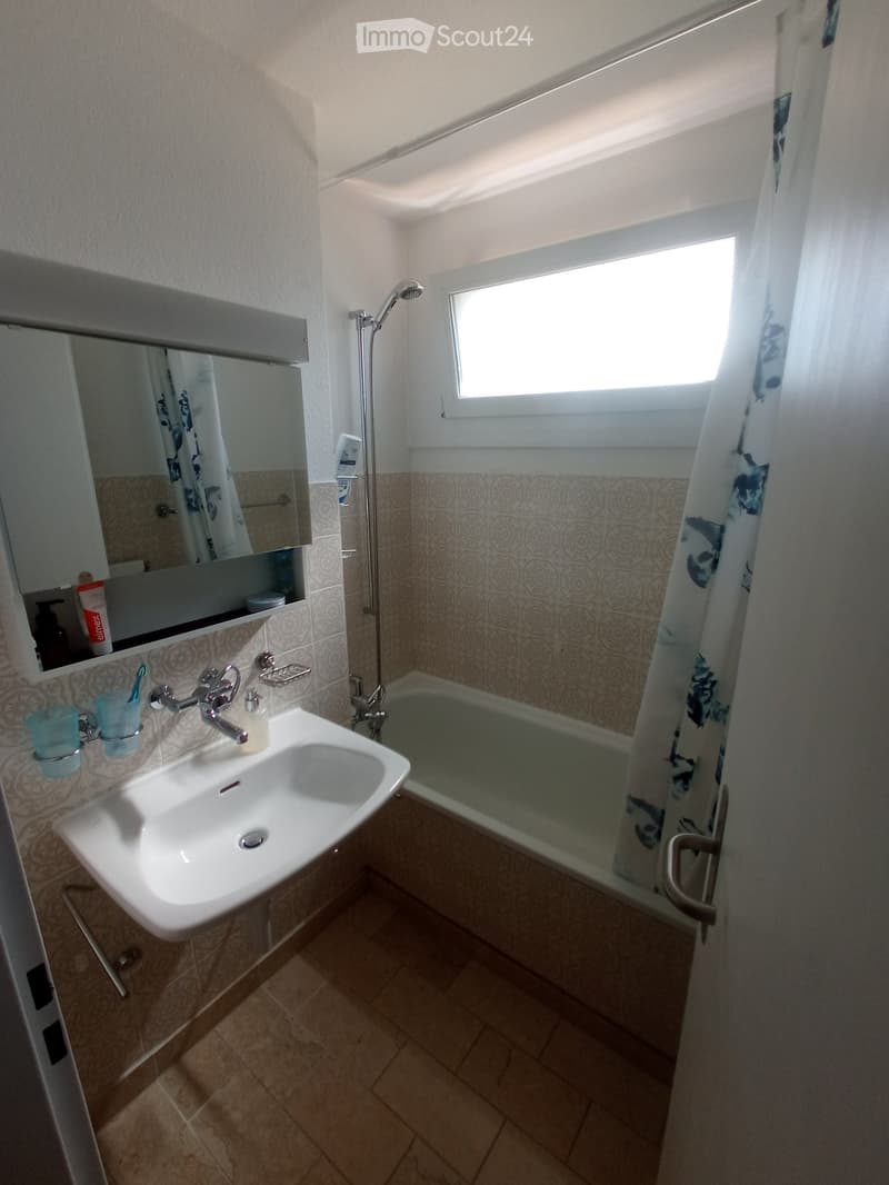 Bathroom with a white sink, mirror, bathtub, and shower. The walls are tiled with a decorative pattern, and the floor is tiled in a warm beige color.