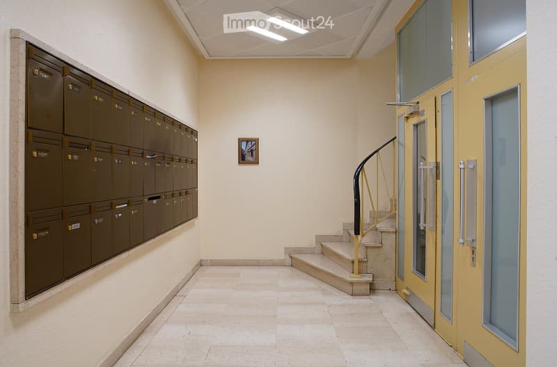 Hallway with beige walls, tiled floor, mailboxes on one side, staircase with yellow handrail, and a framed picture on the wall.