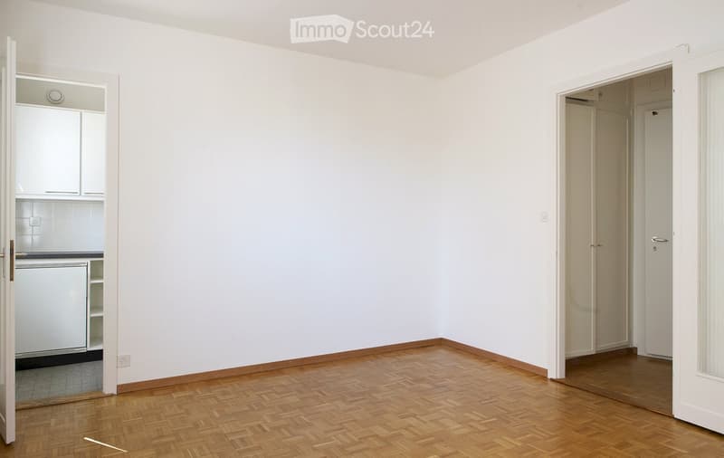 Empty room with wooden flooring and white walls, featuring an open doorway to a kitchen area and another doorway to a hallway.