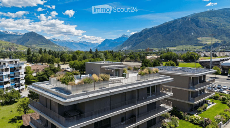 Modern multi-story building with flat roof, solar panels, and rooftop garden overlooking a scenic mountain range.