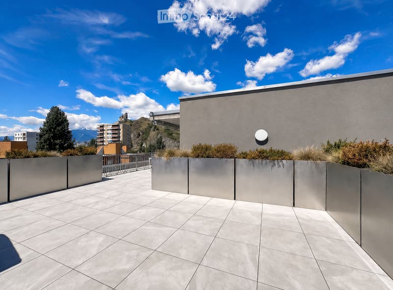 Modern rooftop terrace with grey tiled flooring, metal planters, and a low wall, overlooking a cityscape with mountains in the background.