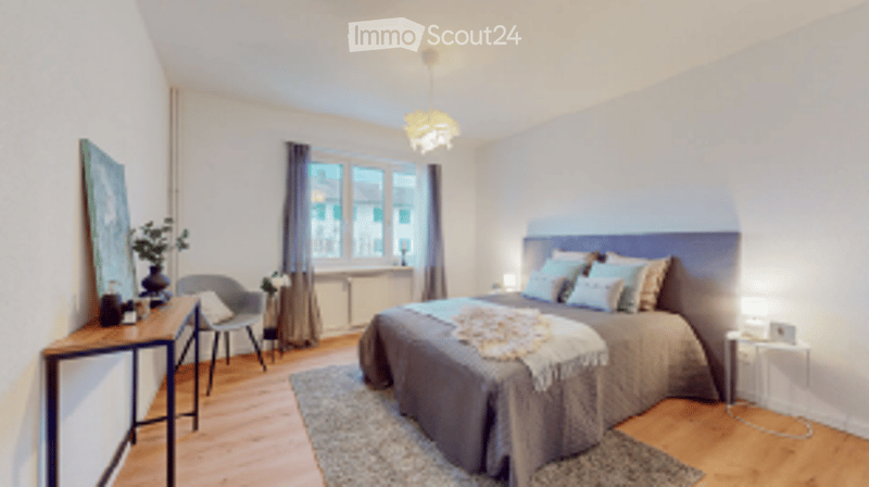 Bright bedroom with wooden flooring, large window, bed with pillows and blanket, nightstand with lamp, small table with chair, and a picture frame on the wall.