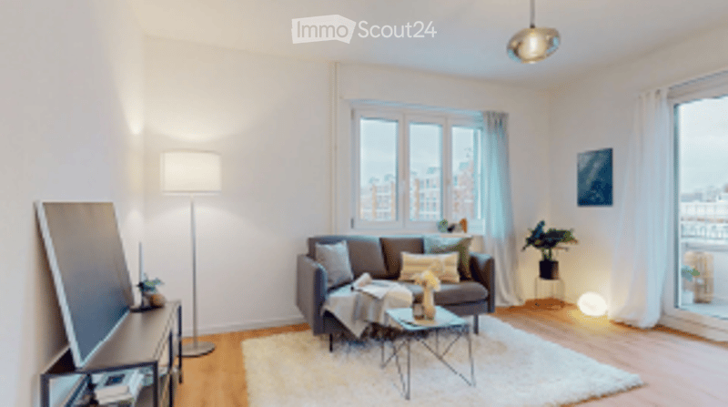 Bright, modern living room with large windows offering a city view. Features a grey sofa, coffee table, floor lamp, side table with a plant, and a mirror on the wall.
