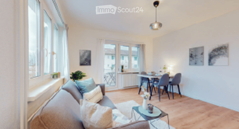 Bright living room with large windows, grey sofa, coffee table, dining area with table and chairs, pendant light, and artwork on the walls.