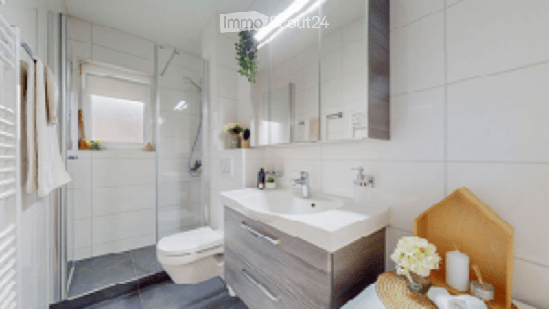Modern bathroom with white tiles, a large mirror, a sink with a modern faucet, a shower, a toilet, and a wooden cabinet. A towel hangs on the left side, and a small plant is placed on the windowsill.