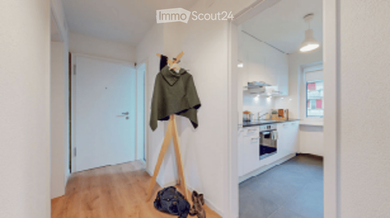 A narrow hallway leads to a modern kitchen area with white cabinets, a built-in oven, and a sink. A coat rack with a jacket and shoes is visible in the hallway.