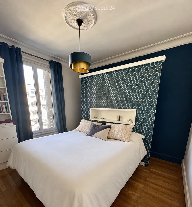 Modern bedroom with a large bed, decorative pillows, a built-in shelf, a hanging lamp, and a window with dark curtains.