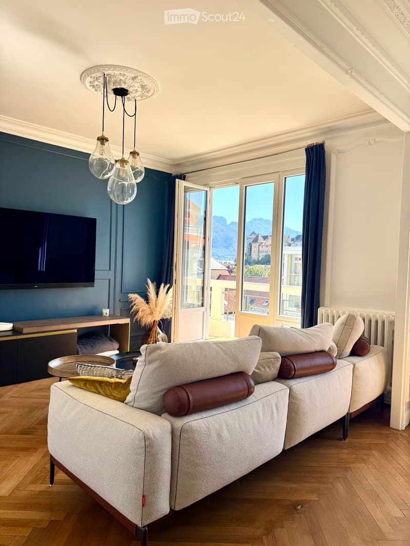 Modern living room with a grey sofa, coffee table, television, and large windows offering a scenic mountain view.