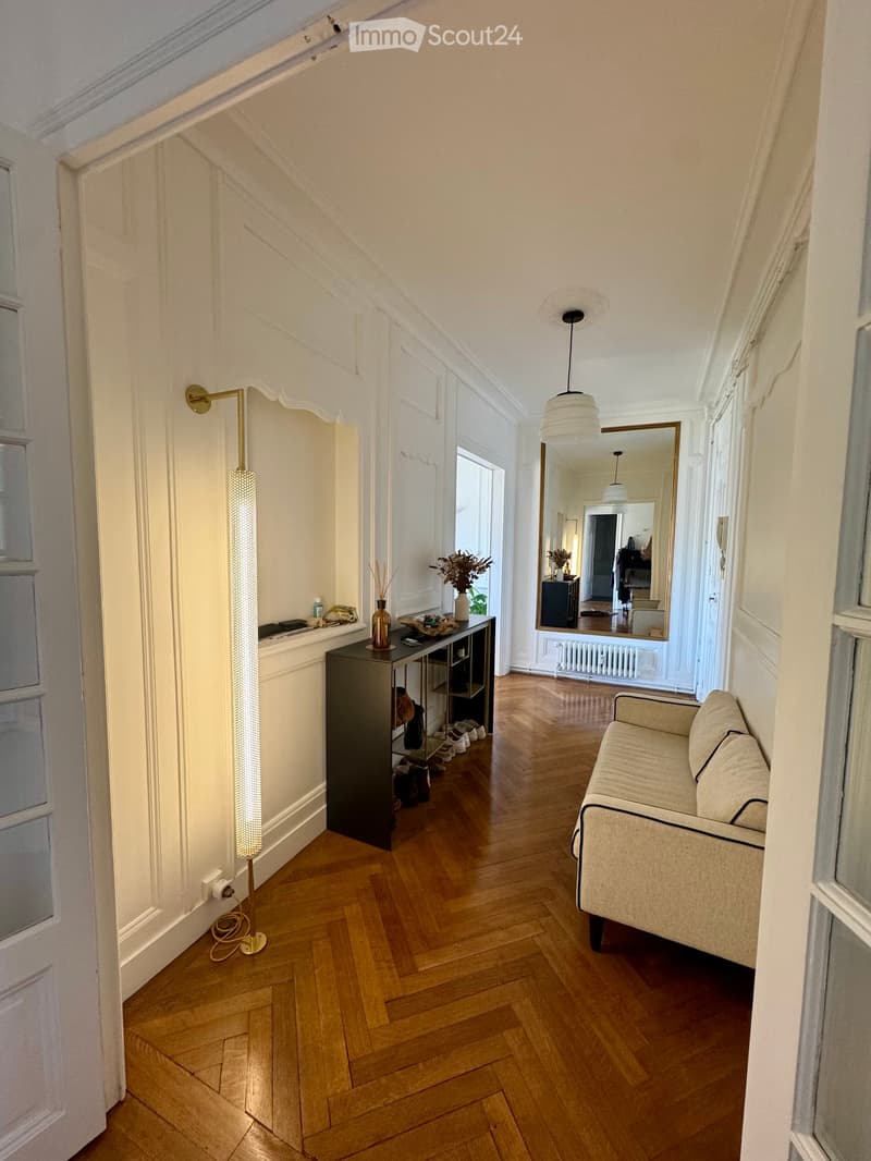 A well-lit hallway with wooden flooring, a sofa on the right, a small table with a vase and decorative items, a tall floor lamp, and a large mirror reflecting part of the room.