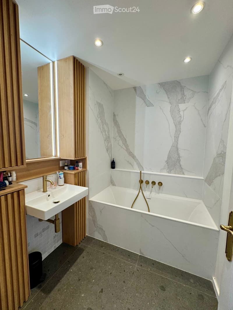 Modern bathroom with a freestanding bathtub, marble walls, a white sink with a gold faucet, and wooden cabinets.