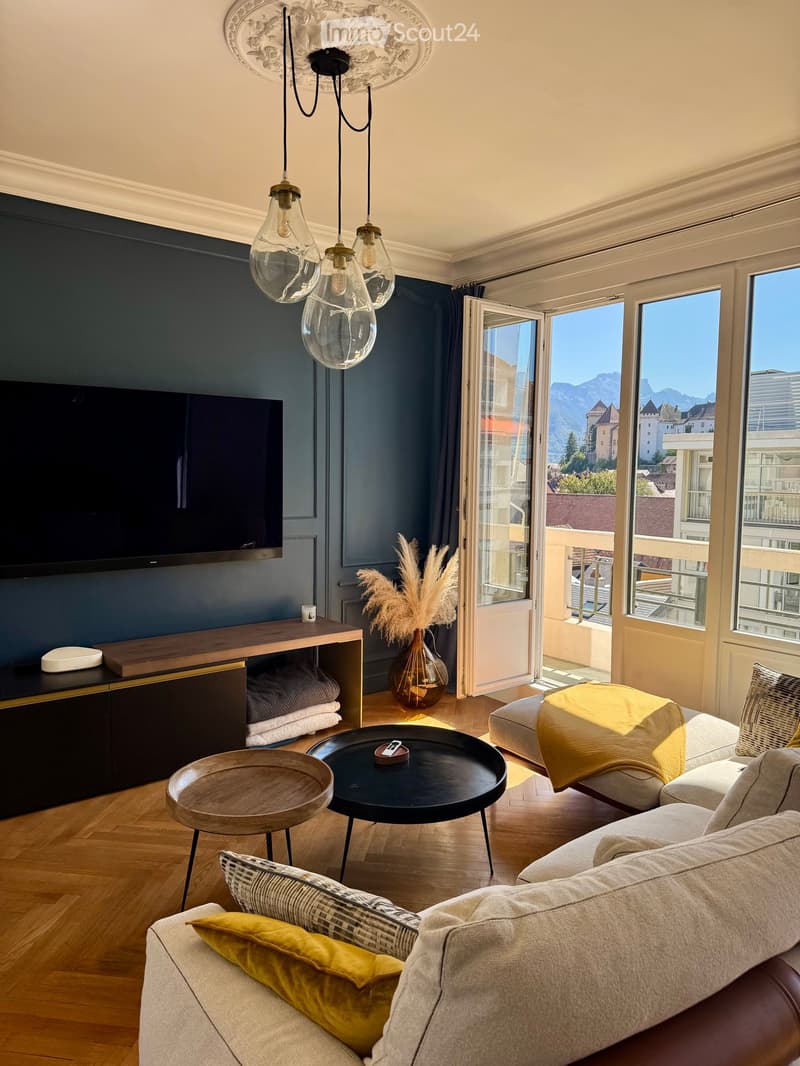Modern living room with a grey sofa, coffee table, side table, television, and pendant lights. The room features large windows with a scenic mountain view and a balcony accessible through glass doors.