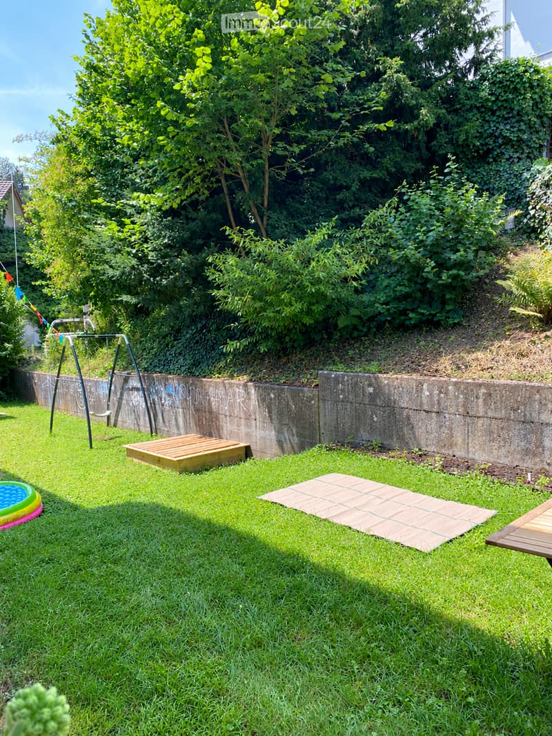 A small, grassy backyard with a swing set, a wooden play structure, a small inflatable pool, a wooden bench, and a raised wooden platform, surrounded by a concrete retaining wall and lush greenery.