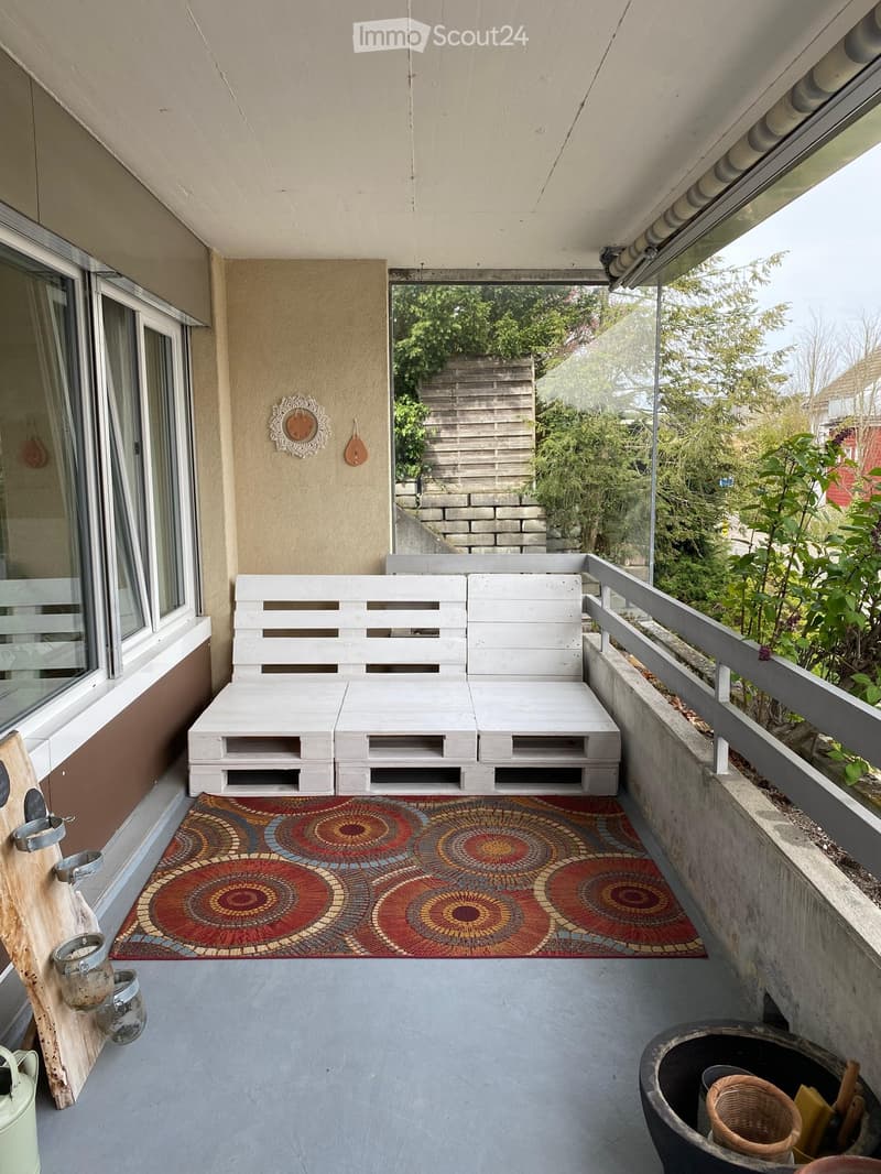 Balcony with a white wooden sofa, colorful circular patterned rug, and decorative wall pieces.