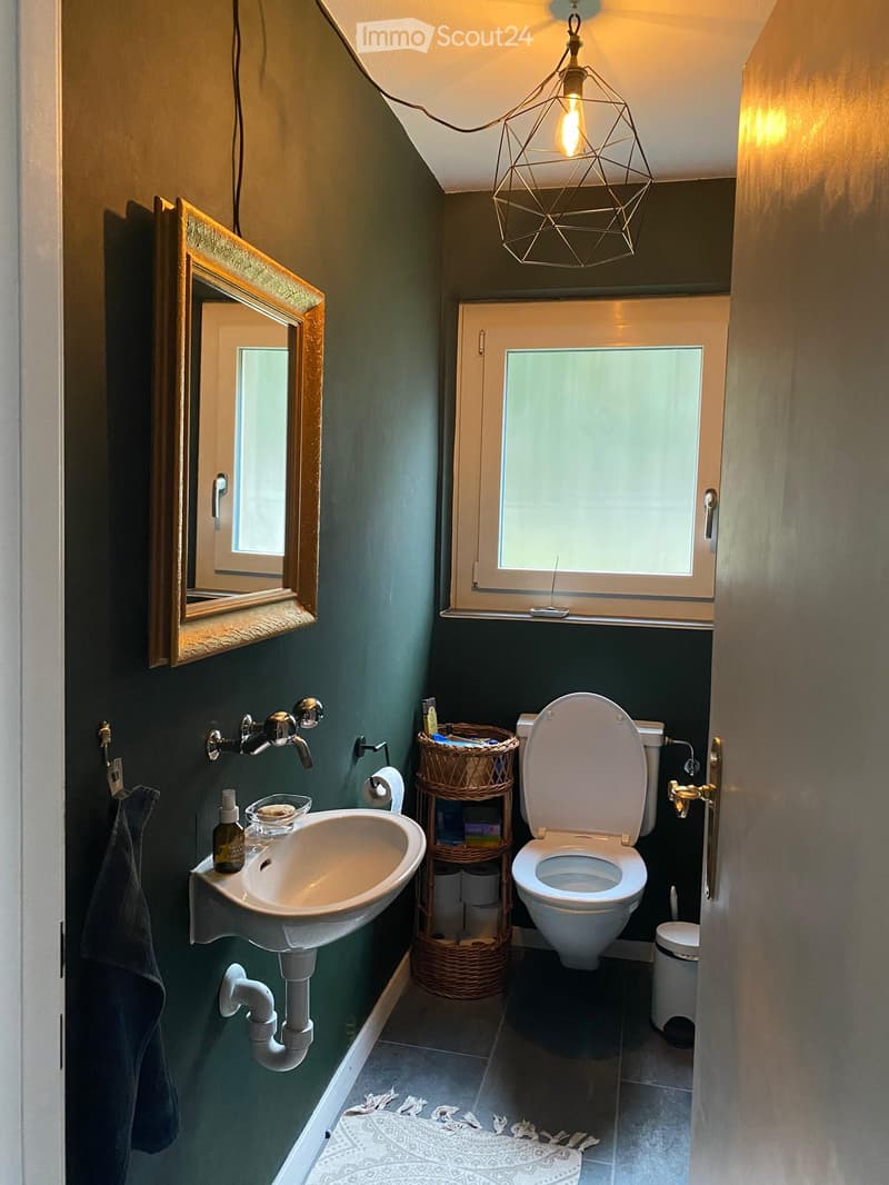 Modern bathroom with dark green walls, white sink, toilet, and a geometric light fixture. Includes a wicker basket on the floor and a mirror on the wall.