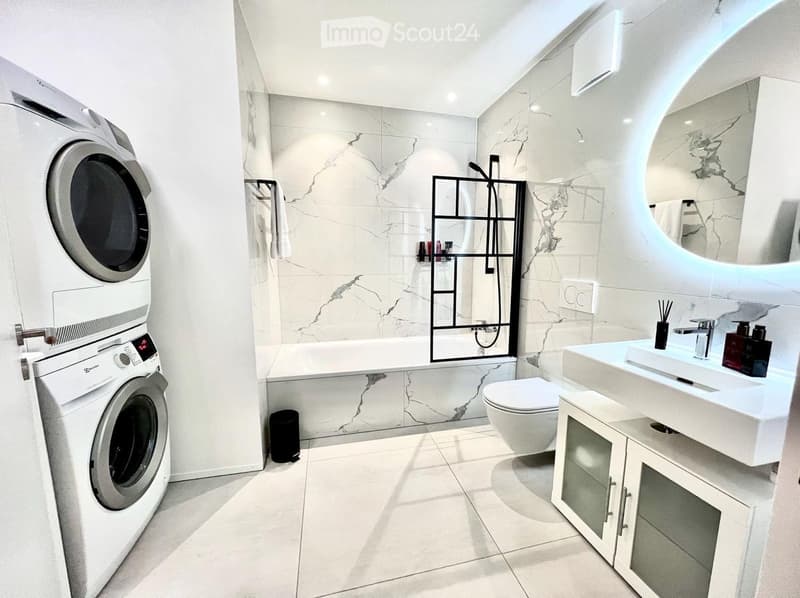 Modern bathroom with marble tiles, a white sink, toilet, bathtub, washing machine, and a large mirror.