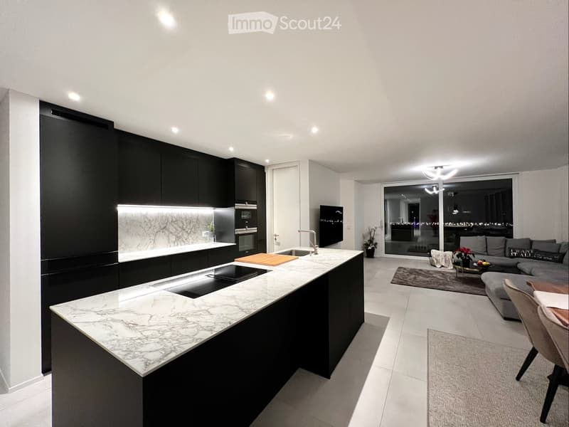 Modern open-plan kitchen and living area with black cabinetry, marble island, grey sofa, coffee table, and dining chair.