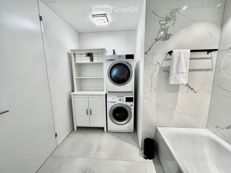 Modern bathroom with a bathtub, white washing machine and dryer, white cabinet, and a towel hanging on a rack.