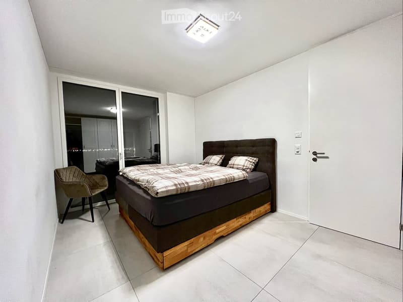 Modern bedroom with a large bed, plaid bedding, a wooden headboard, a single armchair, and large sliding glass doors leading to another room.