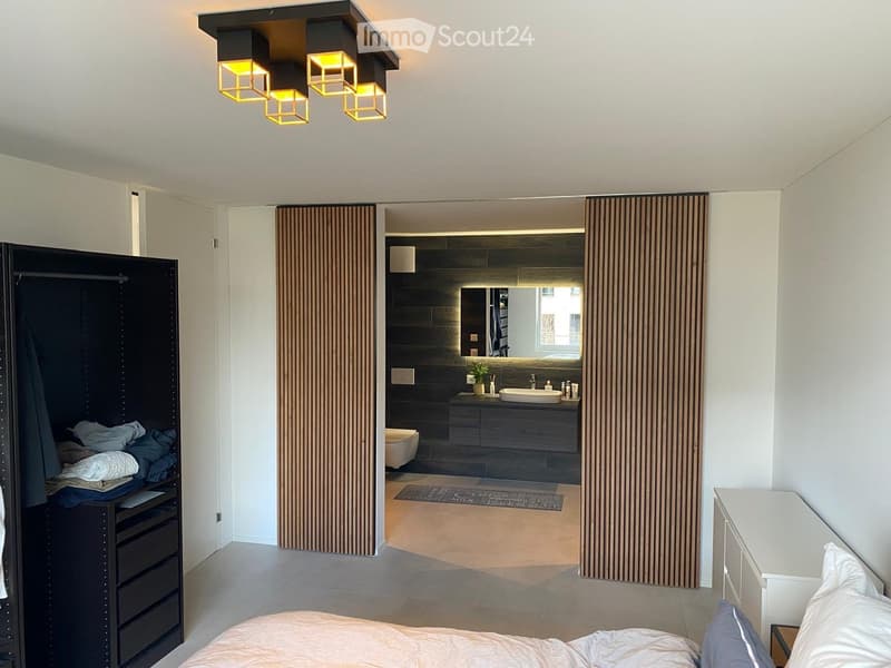 Modern bedroom with a large bed, built-in wardrobe, nightstand, and an adjacent modern bathroom with a sink, toilet, and dark-toned cabinetry.
