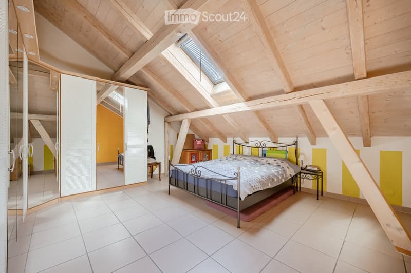 Bright attic bedroom with wooden slanted ceilings, white tiled floor, large window, bed with patterned bedding, nightstand, mirror, and wardrobe.