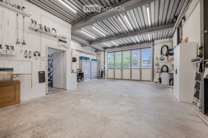 Large, open garage or workshop space with concrete flooring, white walls, exposed metal ceiling beams, and large glass doors leading outside. Shelves hold cleaning supplies, tools, and storage bins.
