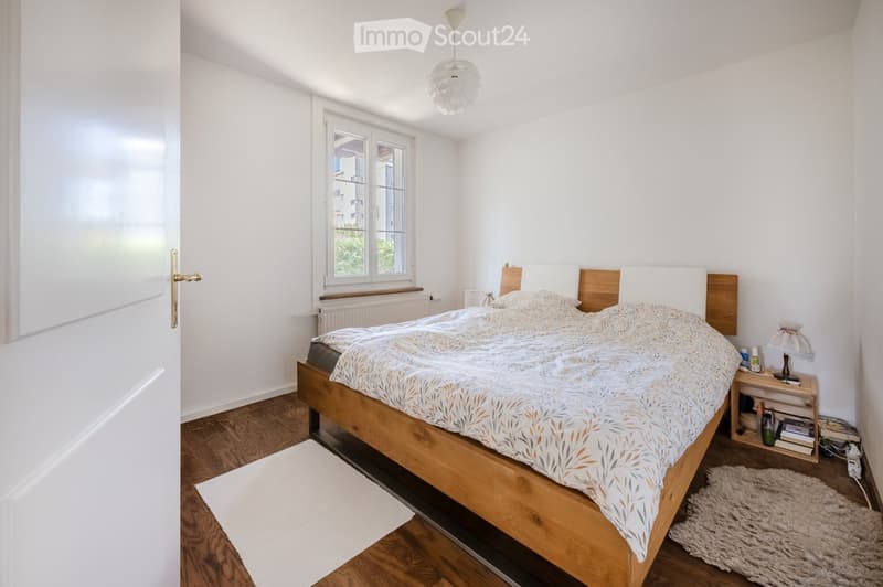 Bright bedroom with wooden flooring, a large bed with a patterned duvet, a nightstand with a lamp, and two rugs on the floor.