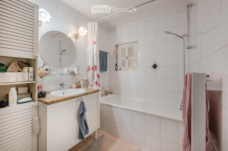 Bathroom with white tiled walls, a sink with a faucet, a bathtub, a mirror, a shower, and a cabinet with various toiletries and towels.