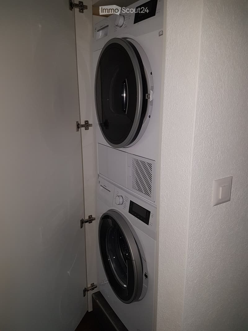 Compact space with a stacked washing machine and dryer unit, white walls, and a door with a latch.