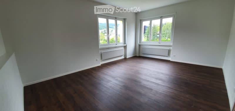 Empty room with wooden flooring, light grey walls, and large windows offering a view of greenery outside. Radiators are visible under the windows.