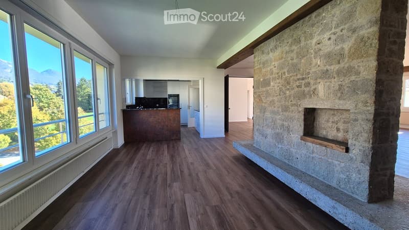 Spacious living area with wooden flooring, large windows offering a scenic mountain view, a stone fireplace, and an open kitchen area with a kitchen island and a mirror.