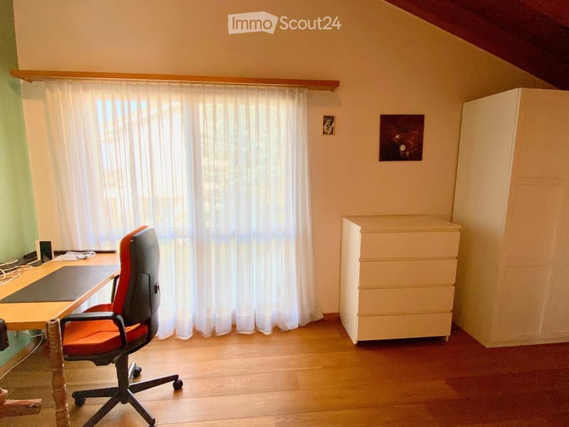 A small, well-lit room with wooden flooring, a large window with white curtains, a red office chair, a wooden desk, and a white cabinet.