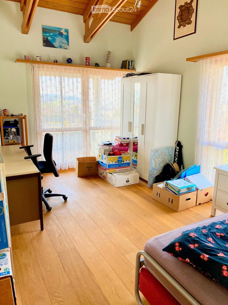 Bright bedroom with wooden flooring, large window with sheer curtains, bed with floral bedding, desk with chair, and various storage boxes and items.