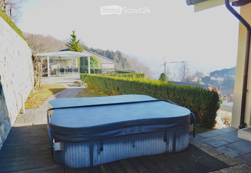 Outdoor area with a wooden deck, hot tub, neatly trimmed hedges, and a scenic view of the surrounding landscape and distant buildings.