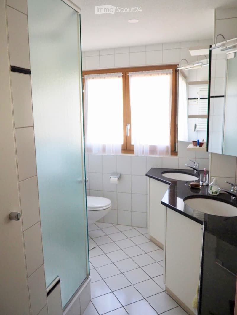 Modern bathroom with white tiled walls and floor, a toilet, a double sink vanity with a black countertop, and a glass shower enclosure.