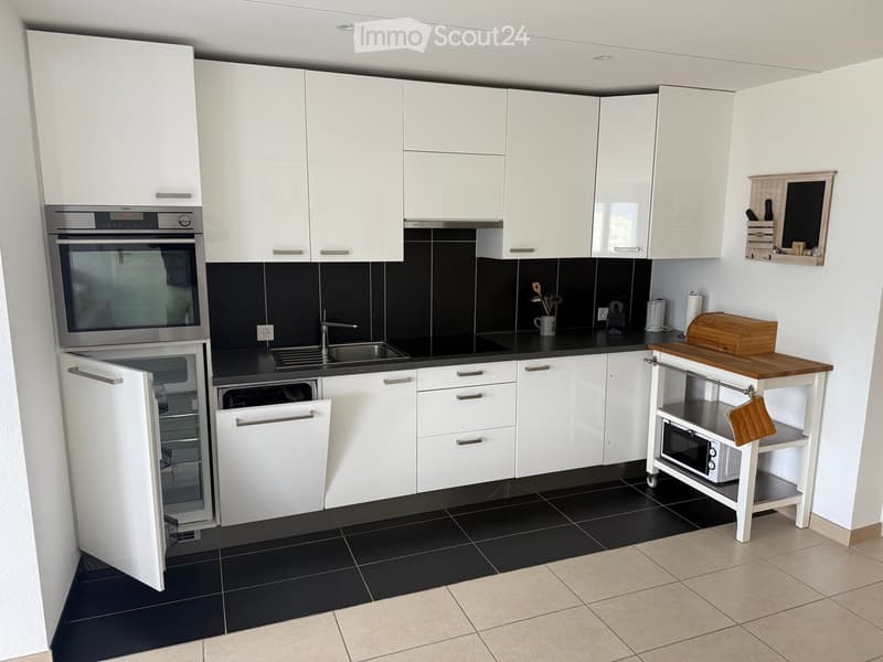 Modern kitchen with white cabinets, stainless steel appliances, black countertops, and a small wooden cart with a microwave and cutting board.